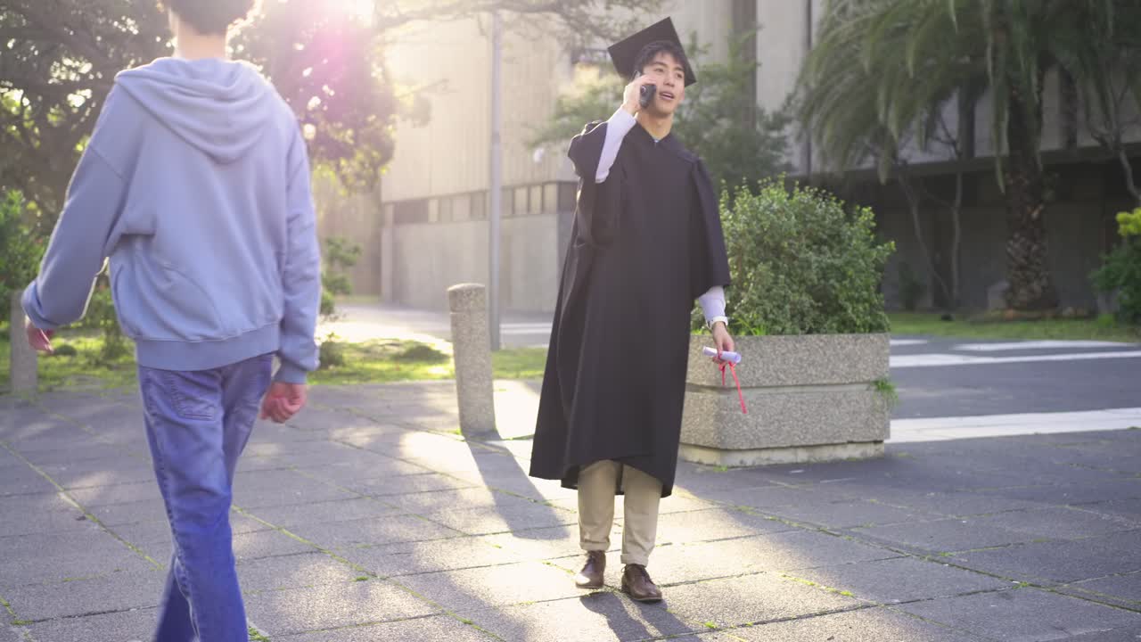 Uomo asiatico in tocco e toga nel campus che tiene il diploma, risponde a una chiamata di congratulazioni sullo smartphone mentre cammina