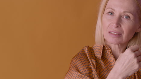 happy blonde mature woman wearing brown shirt dancing, smiling and touching her hair on orange and purple background