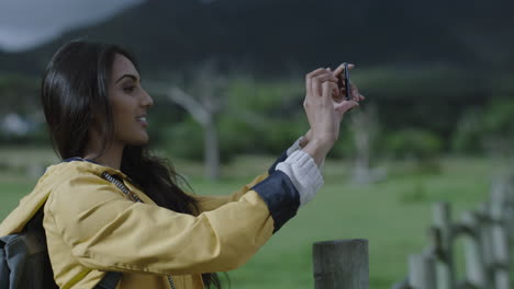 attractive young indian woman taking photo using smartphone camera technology enjoying sharing adventure travel hiker photographing green countryside outdoors on mobile phone