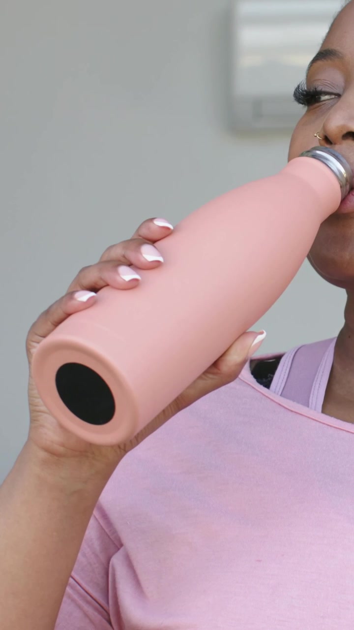 Vertical video: Tilting woman sipping pink bottle at home to hydrate, holding cap, sports-bra strap