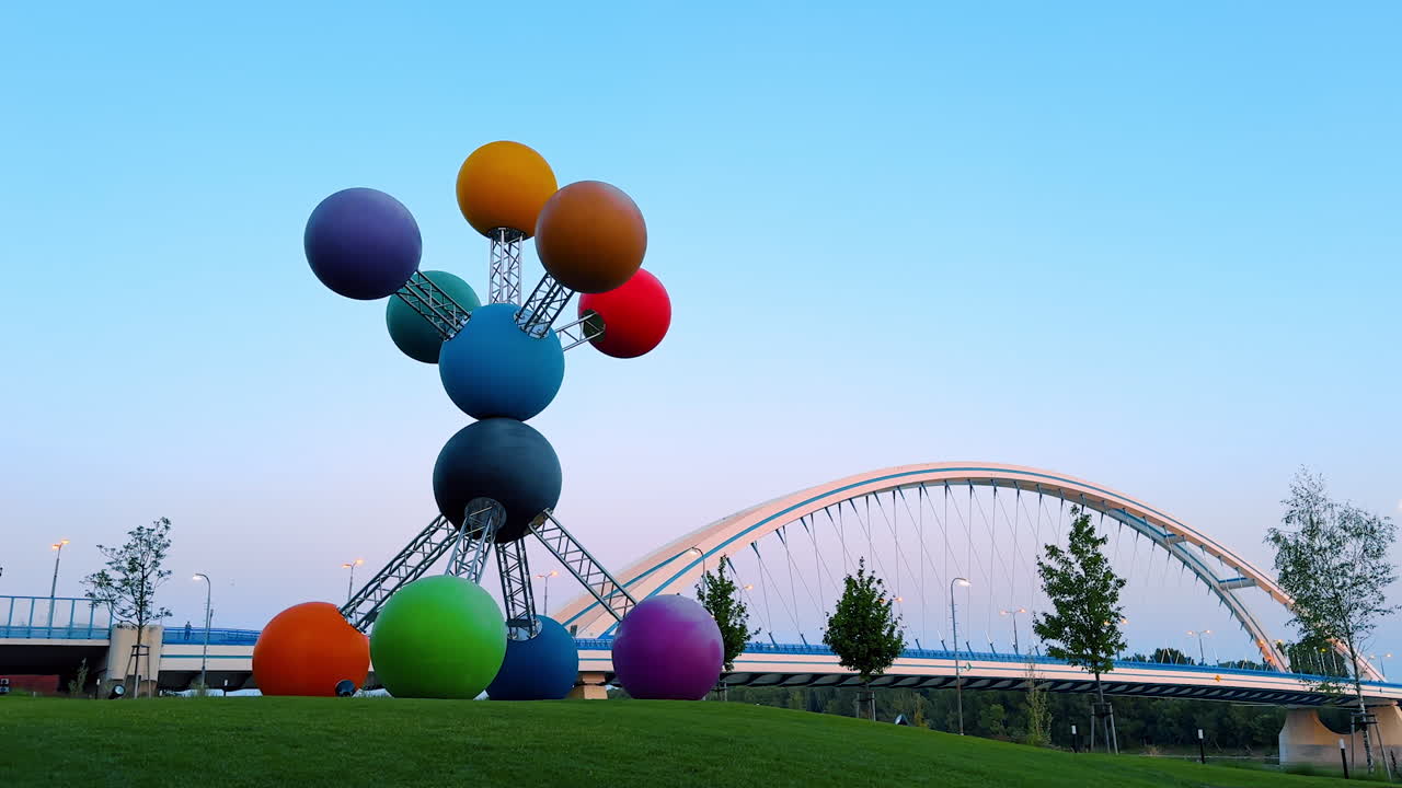 Bratislava, Slovakia, 2 June 2025: Vivid sculpture by bridge in Bratislava. A striking sculpture made of colorful spheres stands on the grass, with a bridge in the background during twilight