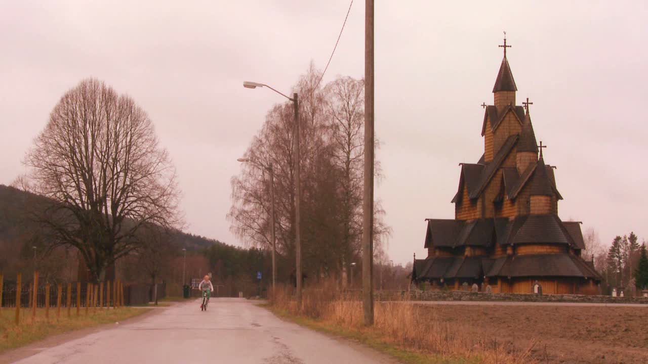Tyttö ajaa pyörällä vanhan puisen sauvakirkon edessä Norjassa