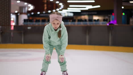 a close view of a child standing on an ice rink, dressed in a mint green outfit and pink headband, with people and ice skating equipment visible in the background