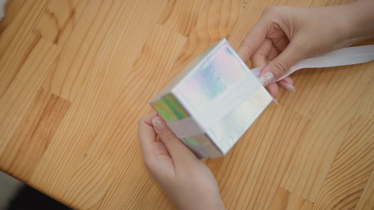 Top-down close-up of two hands finishing gift wrapping by tying satin ribbon around small iridescent square box on pale wooden table, with gentle pulling, smoothing, and knotting in warm indoor light
