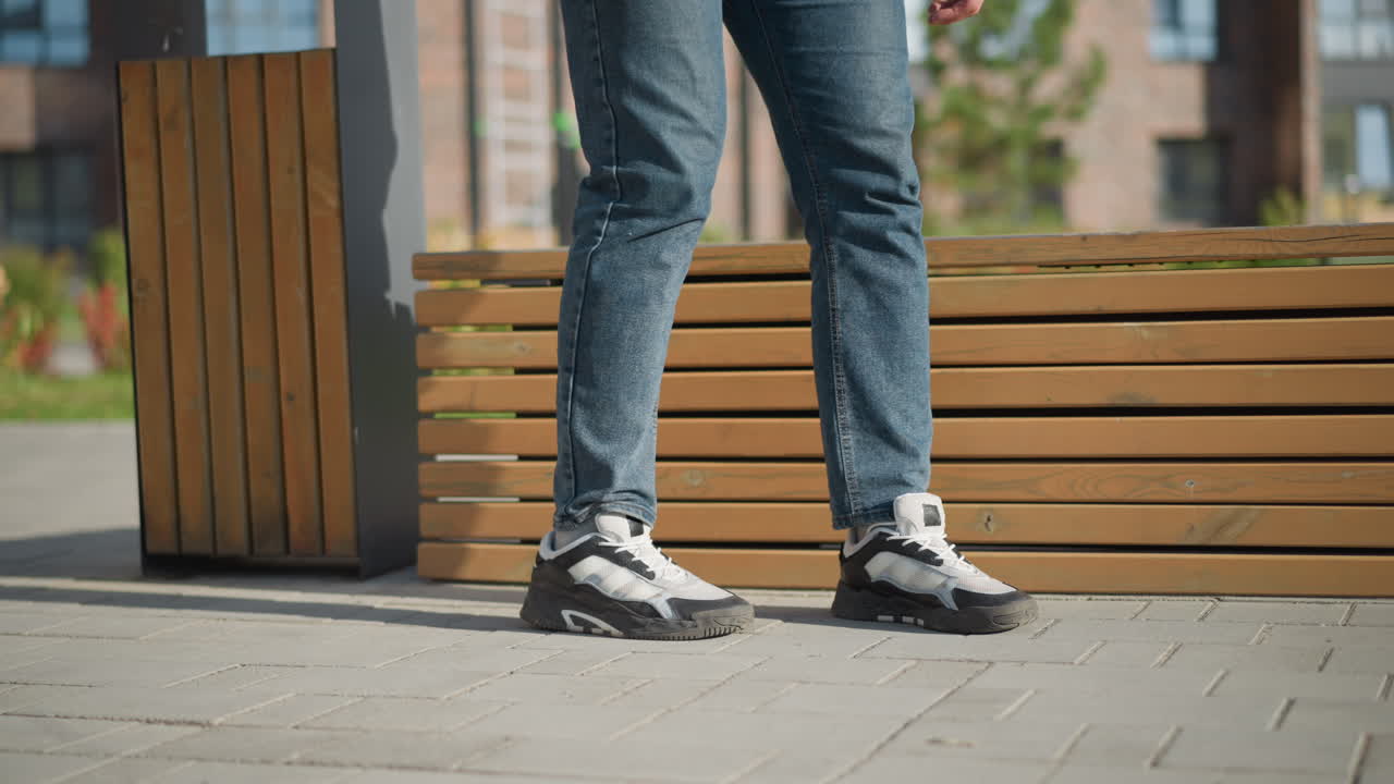 leg view of boy walking toward wooden bench to sit and rest during sunny day in peaceful outdoor setting with visible plants, trash bin, modern buildings, and green lawn in background