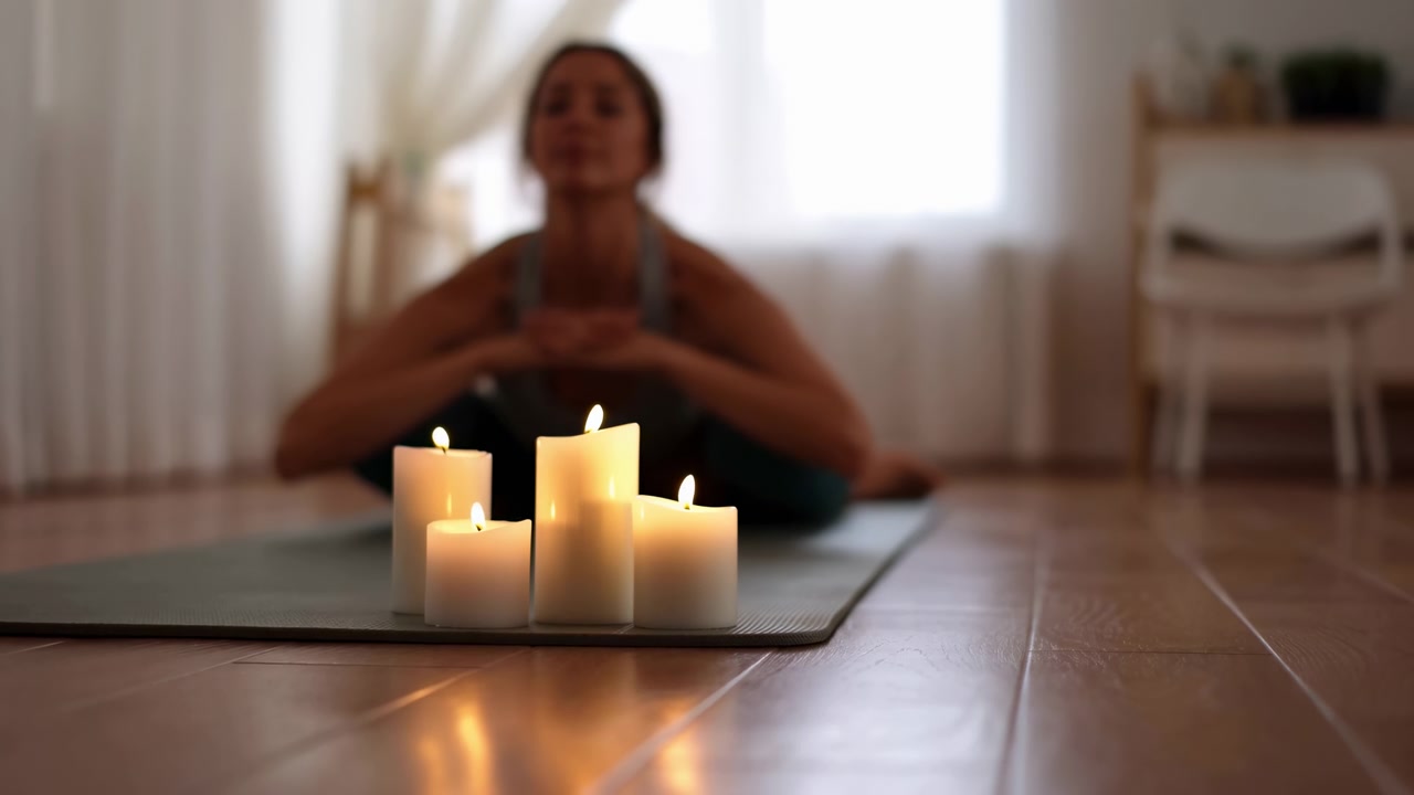 Cinque candele a pilastro accese disposte vicino al bordo di un tappetino da yoga in primo piano, fiamme stabili che proiettano un caldo bagliore ambrato e riflessi su un pavimento di legno lucido, mentre una donna sfocata siede dietro in posizione inclinata in avanti.