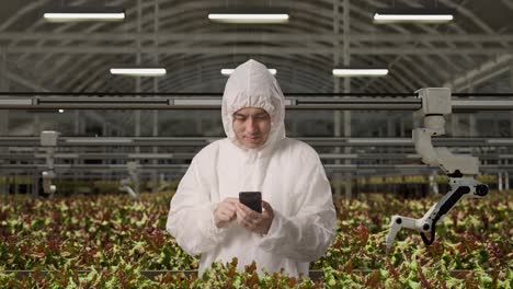 asian man researcher using smartphone while standing in the greenhouse with smart robotic farmers