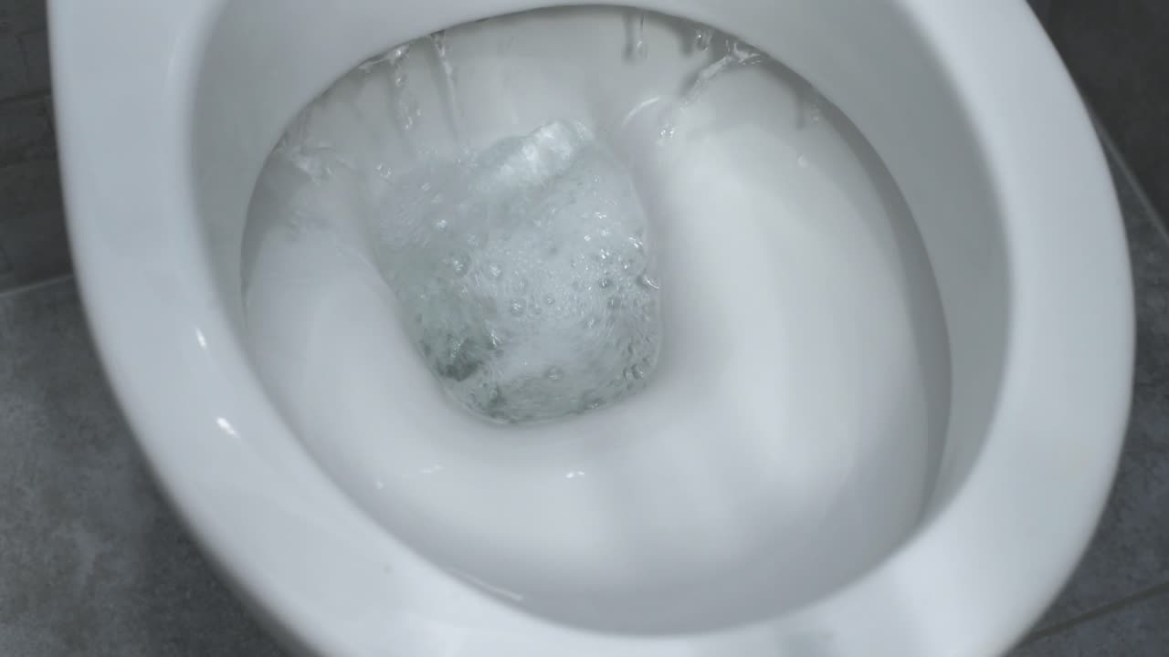 A close-up view of a toilet being flushed, with water swirling rapidly in the bowl. The white porcelain toilet contrasts with the dynamic movement of the water.