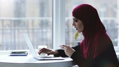 menina muçulmana atraente está digitando algumas informações de um cartão de visita no teclado do laptop. imagens interiores