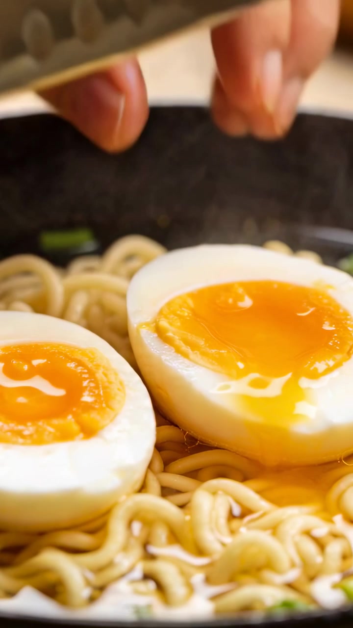 Video vertikal: Tangan menekan dua telur yang sudah dibelah dua dengan pisau koki di atas meja dapur, membiarkan kuning telur melapisi ramen.