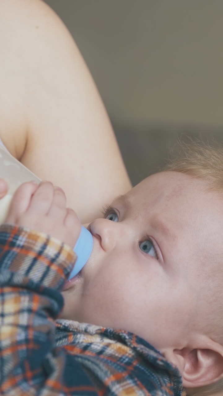 young mommy rocks little son eating milk mix from plastic bottle in spacious light room close view
