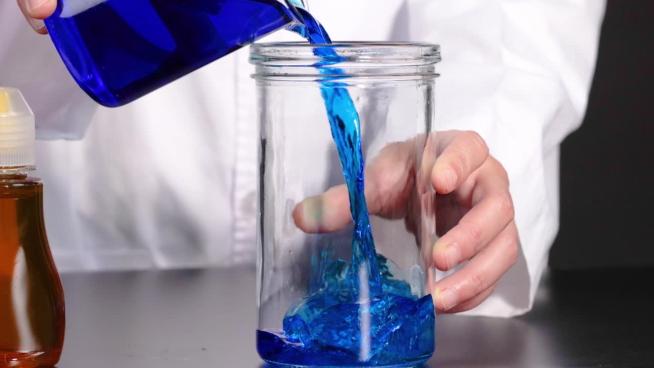 una persona vierte un líquido azul vibrante de una botella en un frasco transparente, mostrando un contraste de color vívido contra un fondo neutral