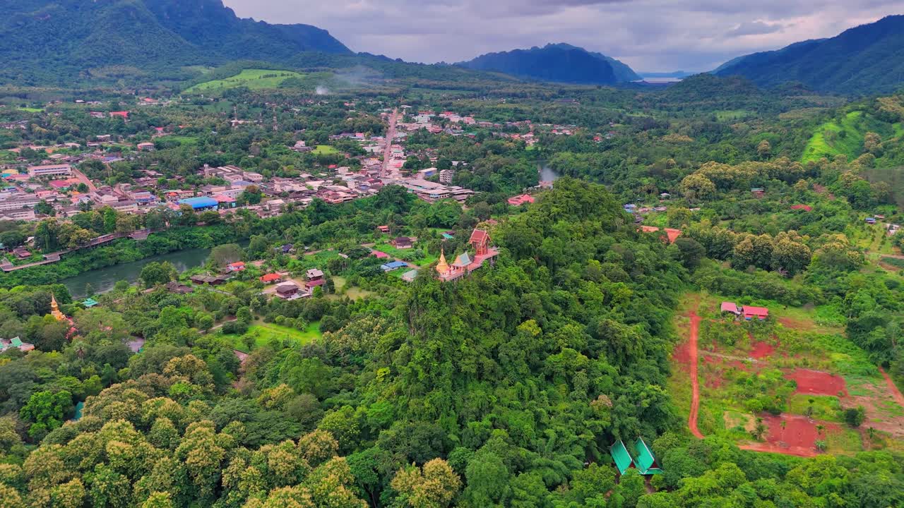 Drone pulls back and ascends from a temple perched on a mountain summit near Sangkhlaburi, Thailand, revealing forest, town, river, and surrounding mountains
