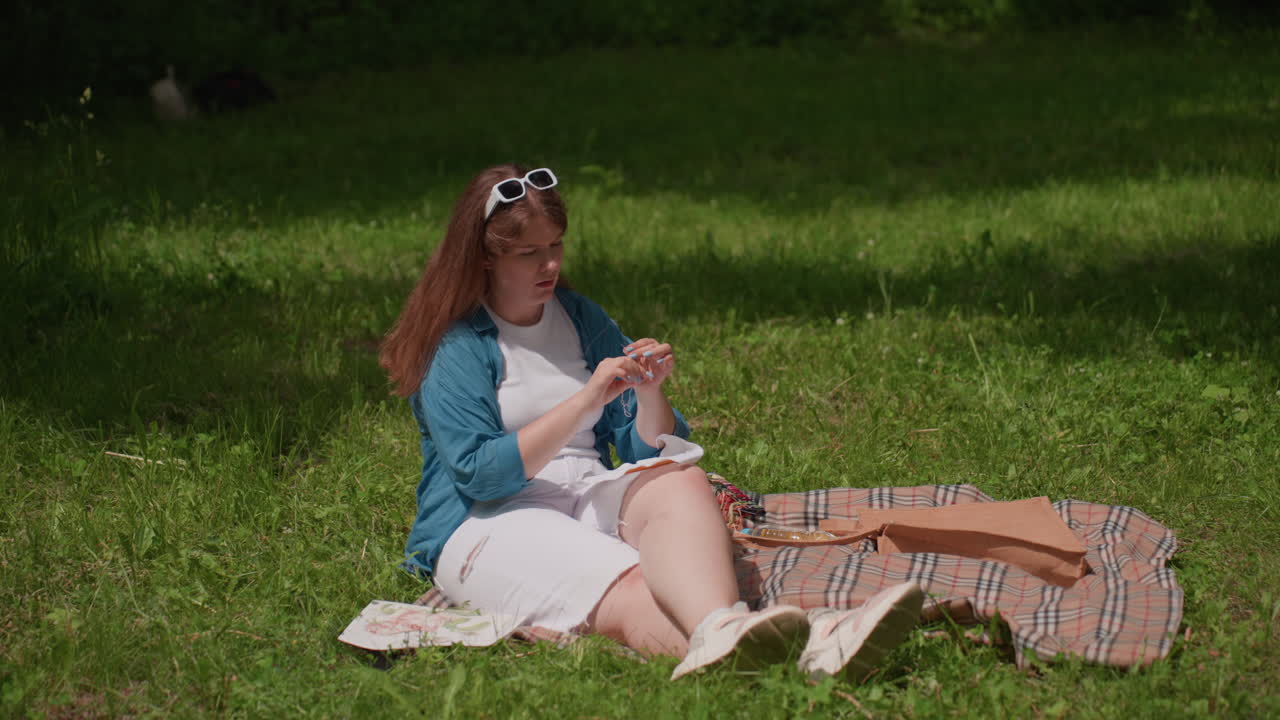 Lady seated on plaid blanket outdoors concentrating on inserting thread into needle eye under bright sunlight, surrounded by soft shadows and green grass, peaceful summer moment of patience