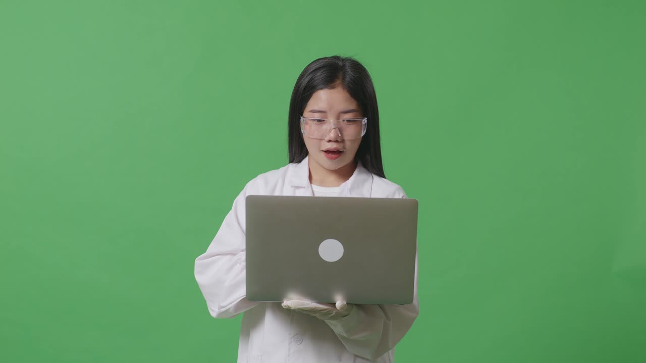 아시아 여성 과학자가 노트북을 보고 비명을 지르며 실험실의 녹색 화면 배경에 서서 축하하고 있습니다.
