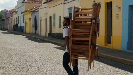 man carrying a large stack of chairs on a cobbled street
