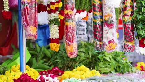 brightly colored flower garlands hang in a vibrant outdoor market stall, surrounded by fresh greenery and blossoms, under natural daylight with slight camera movement