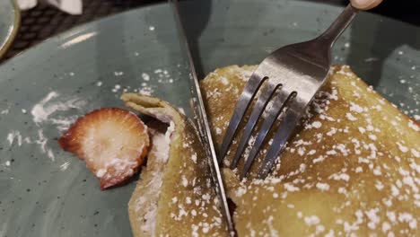 close-up of fork and knife slicing a french crepe filled with cheese cream and fresh strawberries