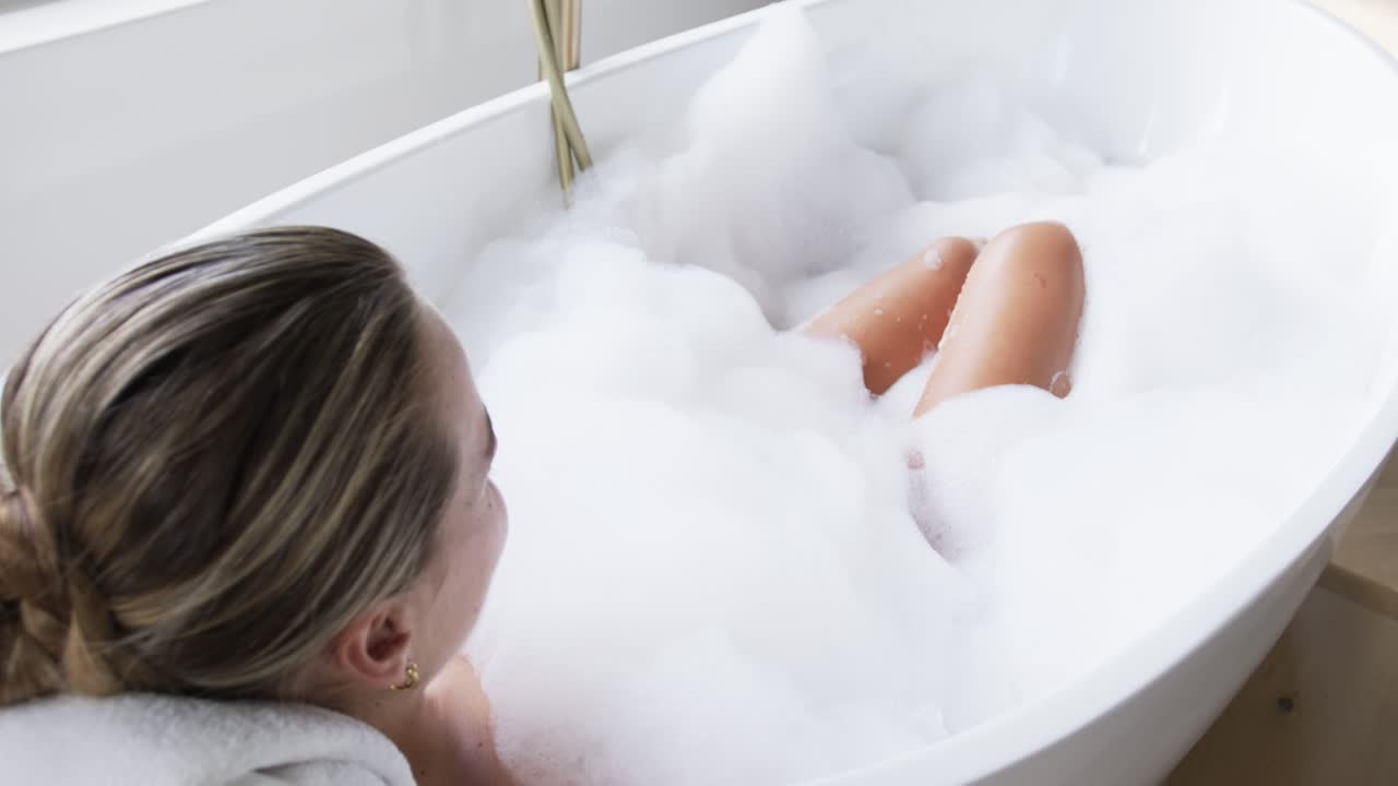Donna sdraiata su un asciugamano nella vasca da bagno, la telecamera esegue una panoramica rivelando una finestra, la schiuma si muove, con spazio per il testo.
