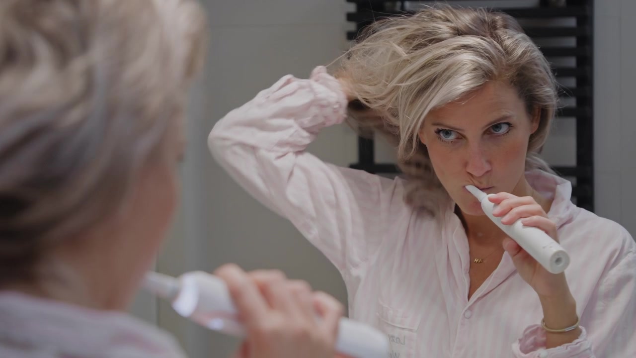 Young caucasian woman checking herself in the mirror while brushing her teeth with an automatic toothbrush in the bathroom. Dental hygiene self-care morning routine.
