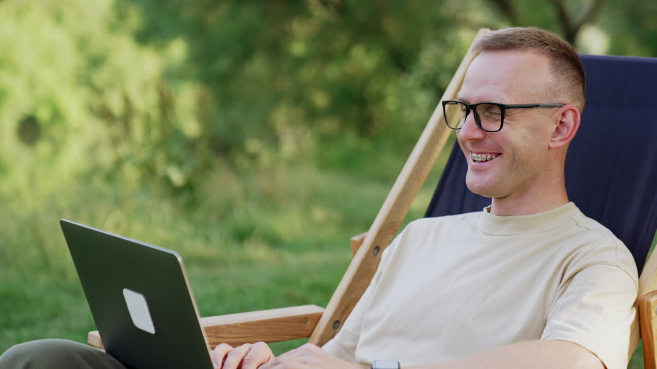 Positive Caucasian man works on laptop in nature. Man in glasses works in a relaxing atmosphere.