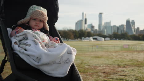 Cute-mixed-race-Asian-baby-sitting-in-stroller-during-winter-in-city-park-with-city-skyline-in-the-background