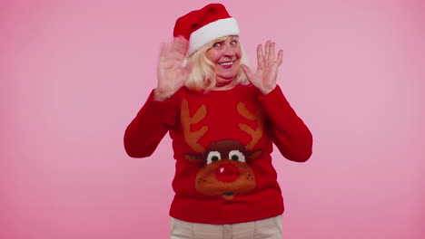 Grandmother-woman-in-Christmas-sweater-with-deers-waves-hand-palm-in-hello-gesture-welcomes-someone