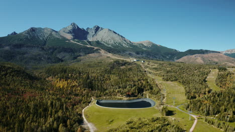 Wide-aerial-drone-shot-moving-from-left-to-right-of-the-High-Tatras-forest-with-a-geomembrane-water-dam-and-cableway,-showcasing-green-summer-trees,-hills,-and-mountains-in-Slovakia,-Europe