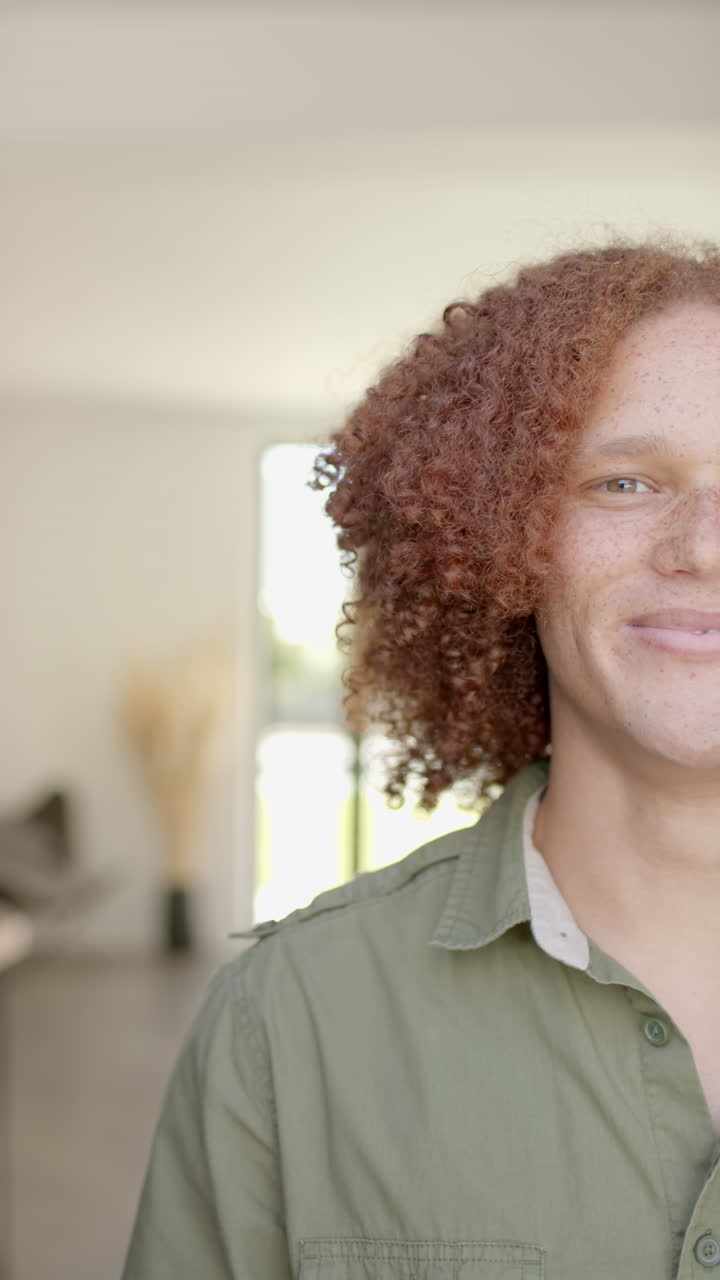 Vertical video of portrait of happy biracial man with curly red hair smiling at home in slow motion