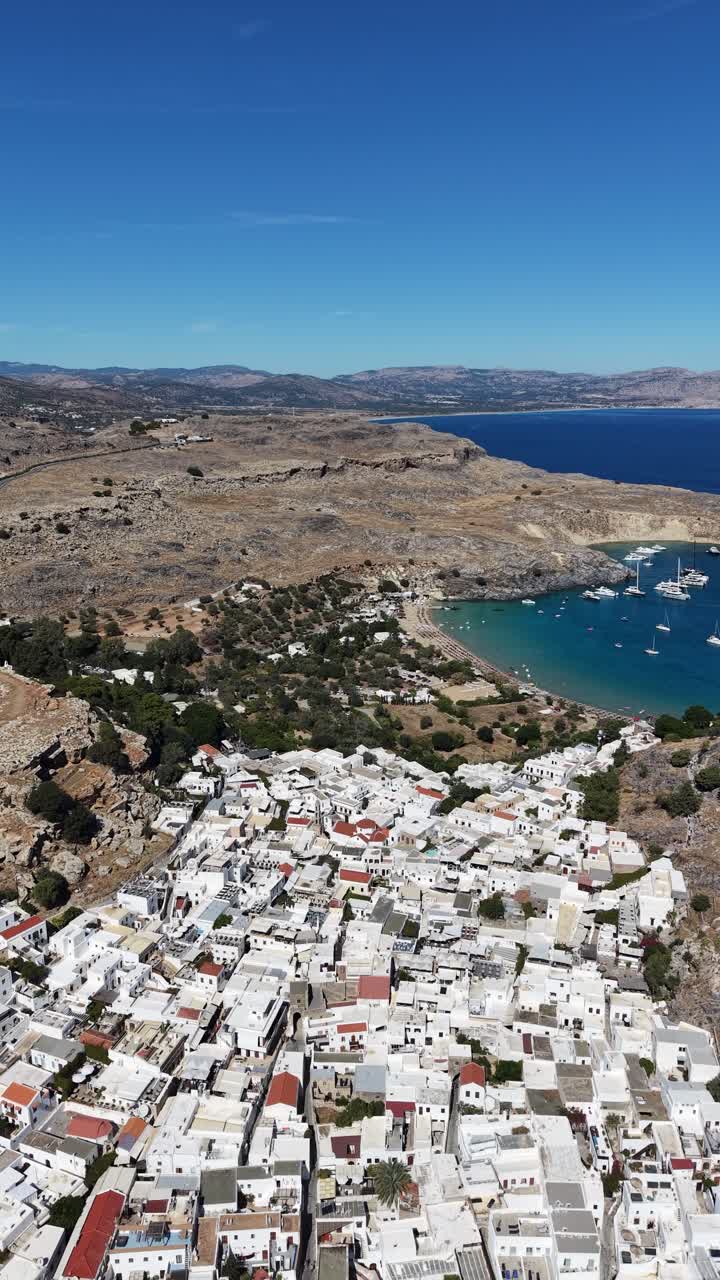Riprese aeree mozzafiato mostrano edifici imbiancati a calce contro un terreno accidentato e il mare blu in Grecia. Questo video verticale offre una prospettiva aerea straordinaria, ideale per la visione su mobile