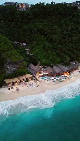 aerial footage reveals a coastal dining spot on a white sand beach in uluwatu, surrounded by clear blue ocean and a dramatic towering cliff that completes the tropical setting.