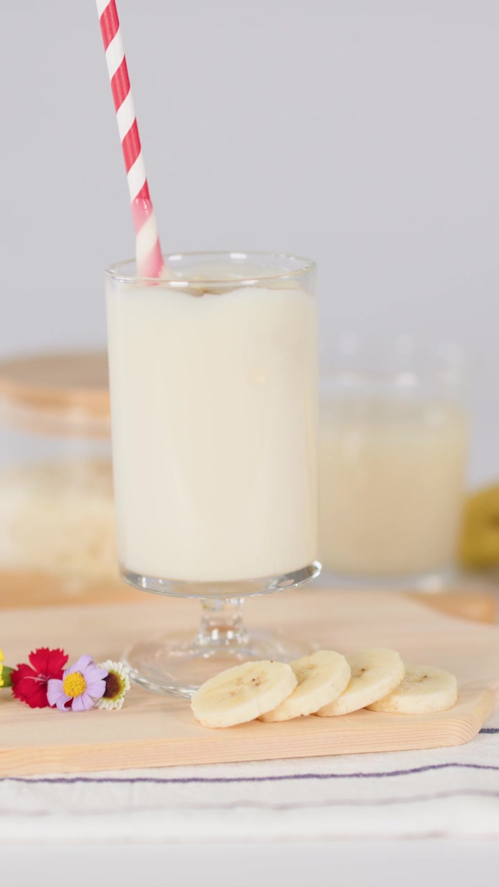 Una mano revuelve un batido cremoso de plátano en un vaso con pajita a rayas, con un estilismo de comida brillante sobre la mesa.