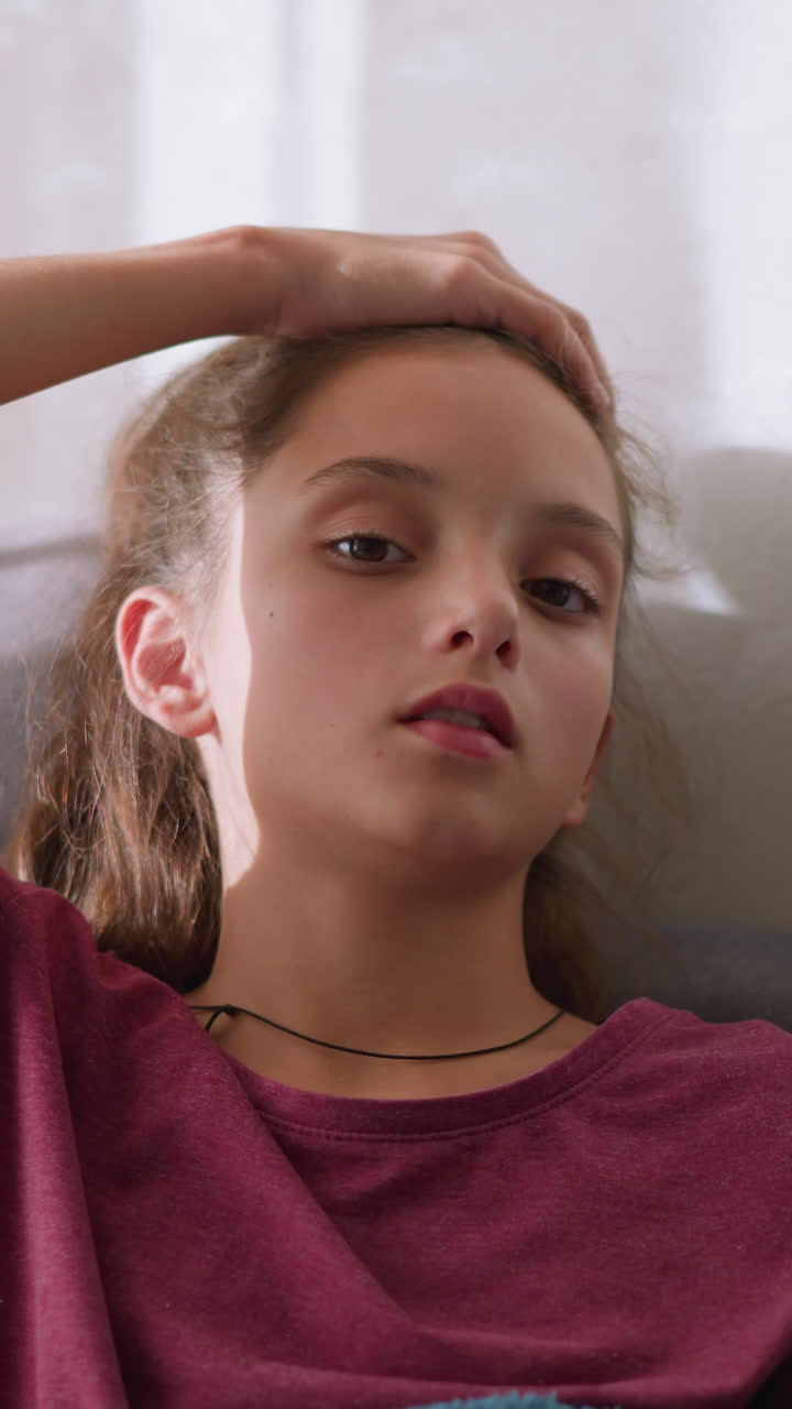 Ragazza pensierosa sdraiata sul divano, mano sulla testa, con un aspetto sofferente e indisposto, persa nei suoi pensieri mentre affronta i sintomi del raffreddore o dell'influenza, che mostra segni di malessere e affaticamento