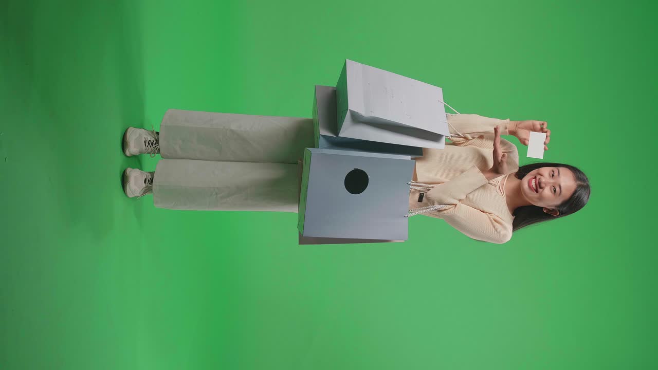 vista completa de una mujer de compras asiática con bolsas de compras sosteniendo una postura de tarjeta blanca y sonriendo a la cámara mientras está de pie frente a la pantalla verde