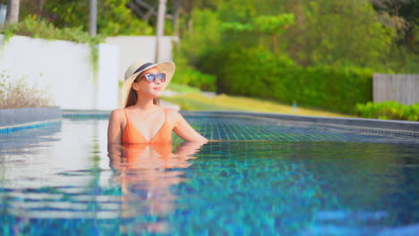 portrait of attractive asian woman relaxing inside tropical hotel swimming pool water wearing orange swimsuit, sunglasses and beach hat in phuket thailand, bust shot slow motion