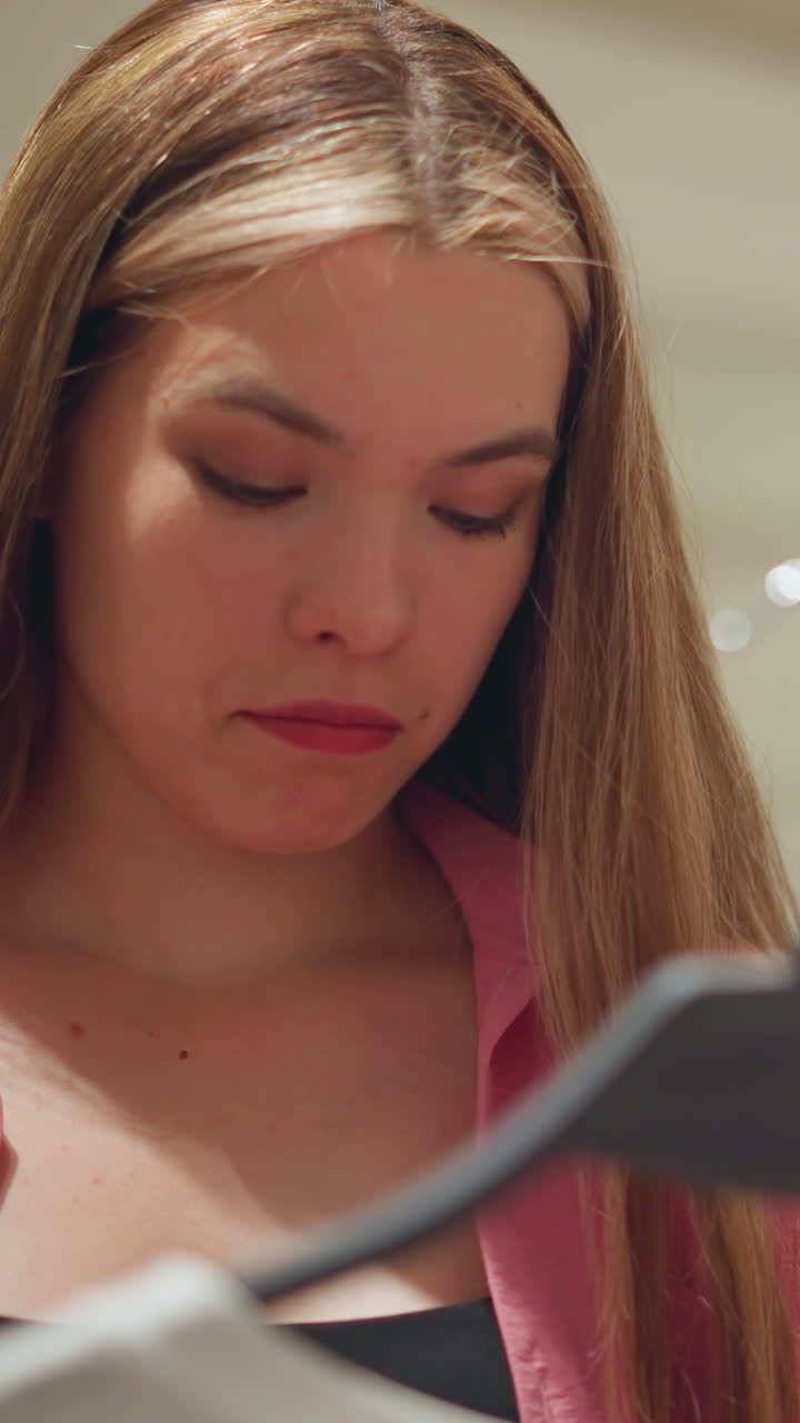 Young lady wearing an unbuttoned pink top thoughtfully evaluates a piece of clothing with an expressive eye movement, other clothes are neatly arranged in the background on a rack