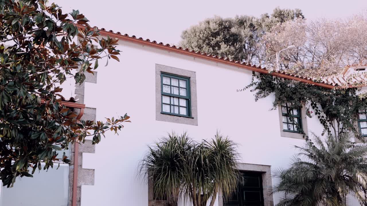 White traditional house with green window frames, tiled roof, and lush surrounding plants