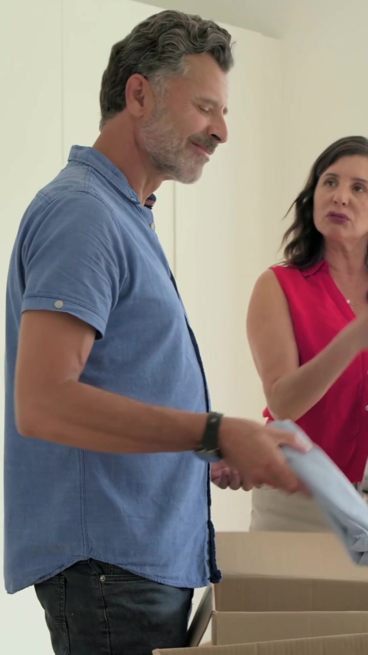 Video verticale: Uomo con camicia blu che disfa i bagagli, passando camicie da una scatola mentre lei organizza l'armadio.