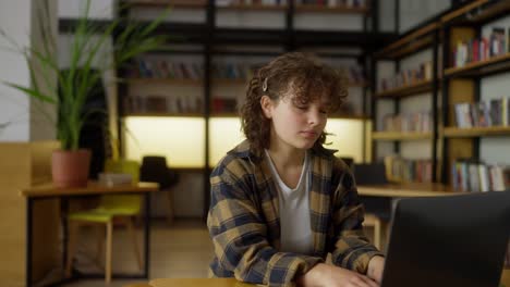 ragazza felice con i capelli ricci che si scalda mentre lavora su un portatile in una biblioteca universitaria