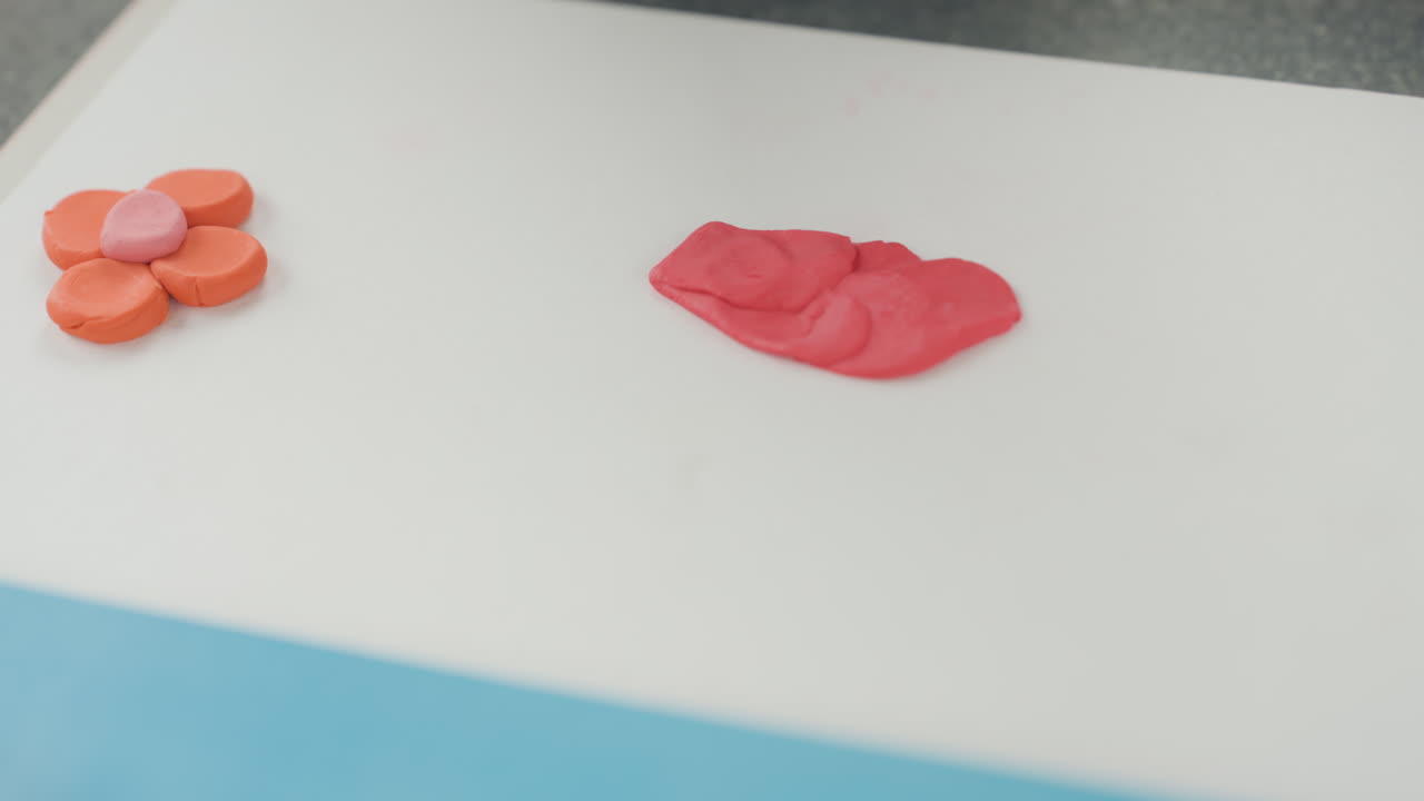 Mãos de criança moldando argila para artesanato de volta às aulas, pequenas pétalas laranja e uma placa vermelha achatada em uma tábua branca, pequena ferramenta preta pressionando o centro, close-up com luz natural suave, estudante preparando arte escolar