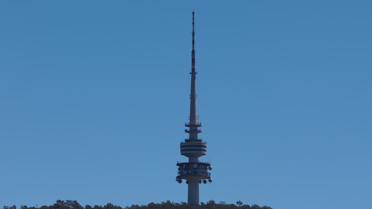 Inquadratura statica e ampia di un'alta torre di comunicazione in cima a una collina, con un cielo azzurro sullo sfondo
