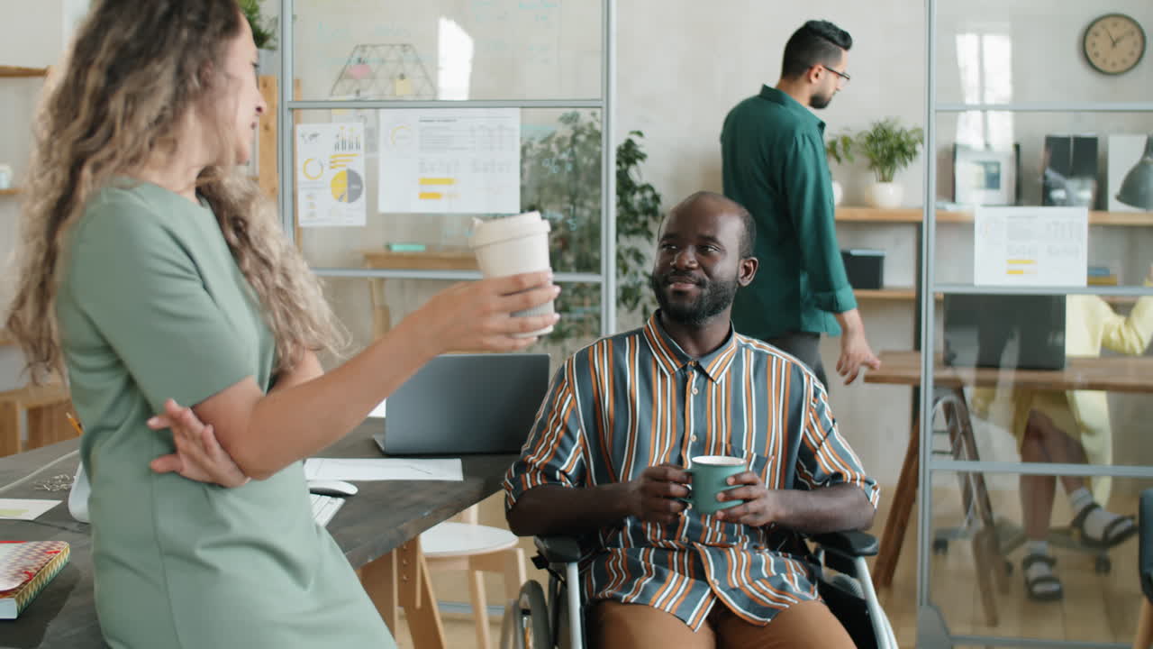 African American Man in Wheelchair Chatting with Female Colleague over Coffee in Office