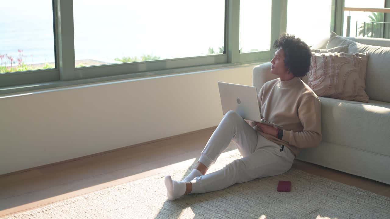African American woman pausing after typing on laptop on rug at home while gazing through window