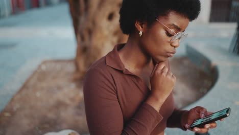 young dark-skinned woman reading news on mobile phone outdoors