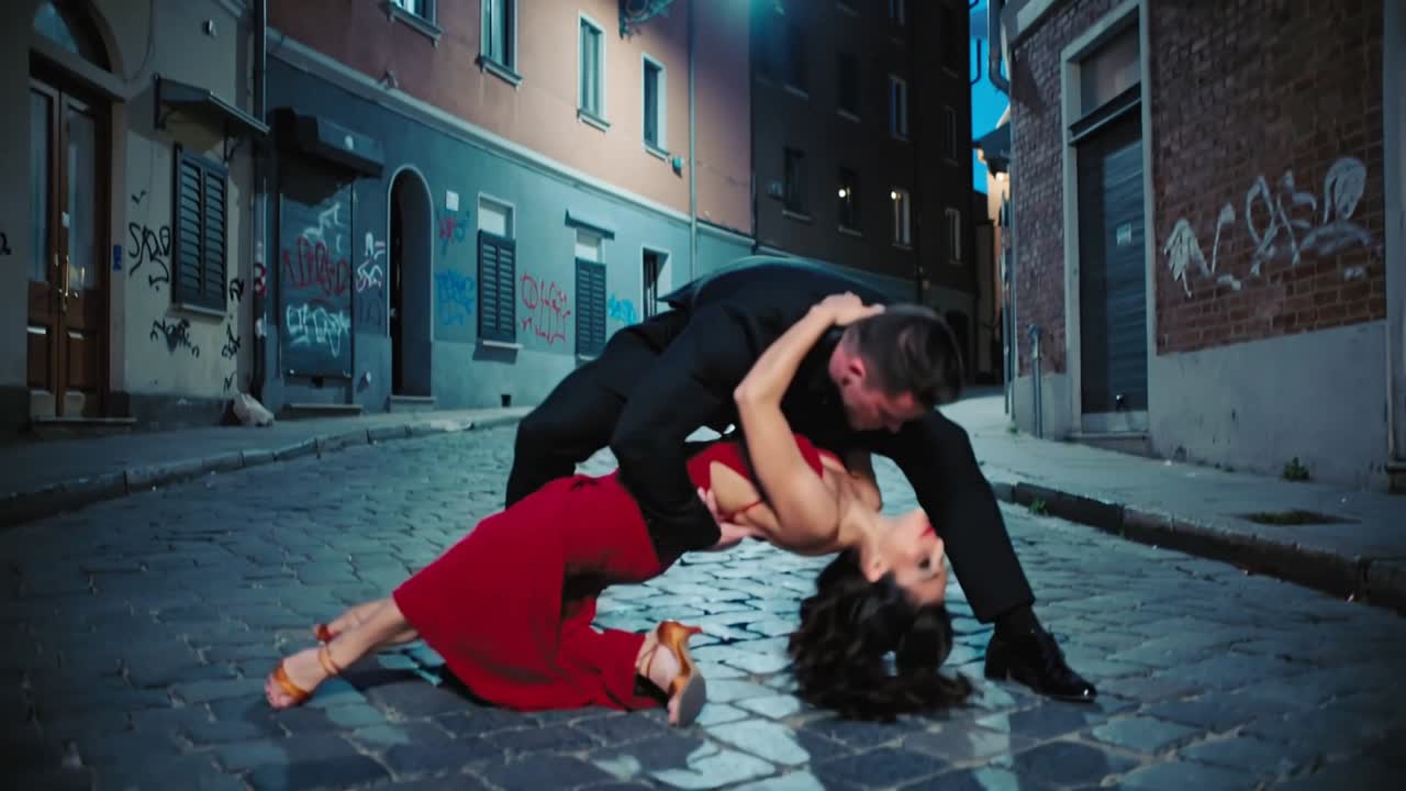 Video verticale: Un uomo guida una donna in abito rosso a fare passi, piroettare, sollevarla e farla scendere sui ciottoli, ballando.