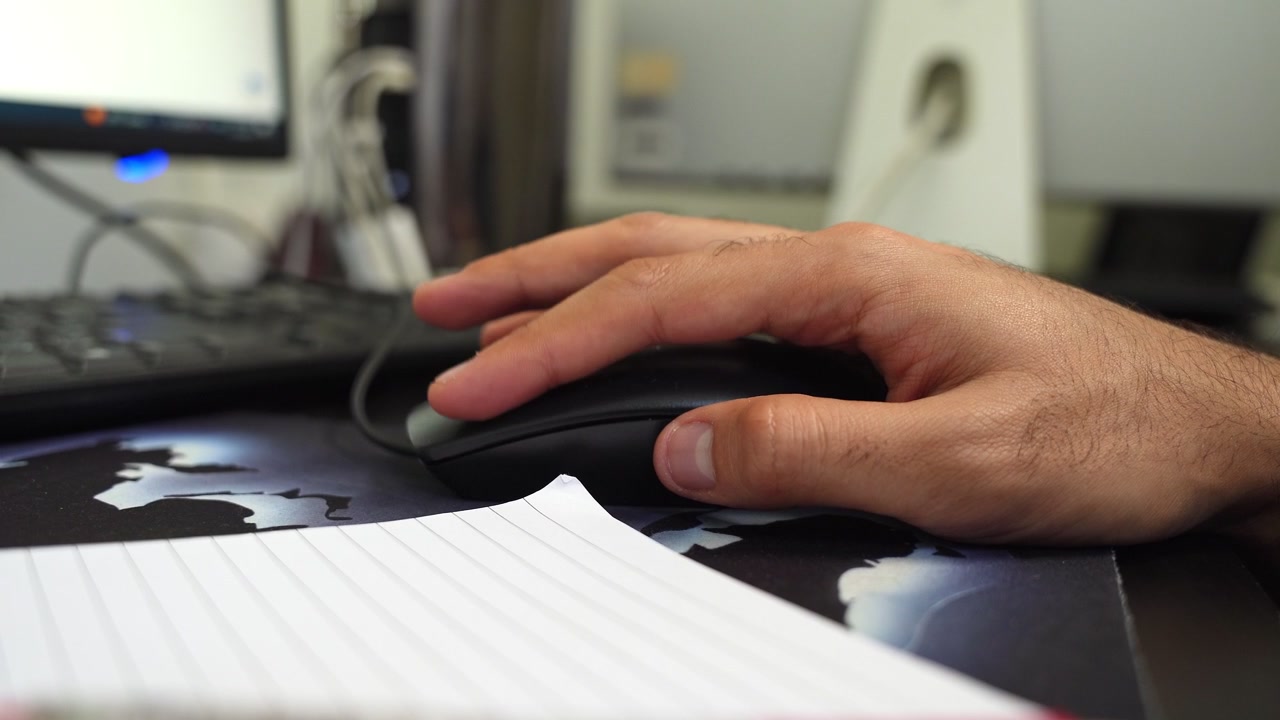 Close-up of a man's hand navigating with a black computer mouse on a desk in a home office setting. Features a keyboard and monitor in soft focus, representing productivity and digital work.