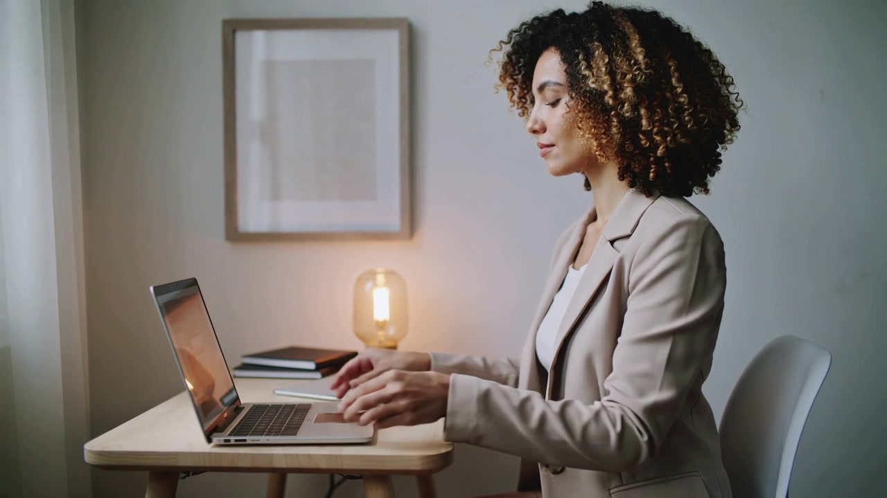 Una donna in blazer beige si avvicina alla scrivania, sistemando il laptop sulla scrivania di legno per iniziare a lavorare.