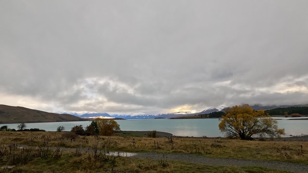 A vehicle moves along a lakeside road at Lake Tekapo, New Zealand, with dramatic clouds, autumn trees, and turquoise water in a wide landscape shot