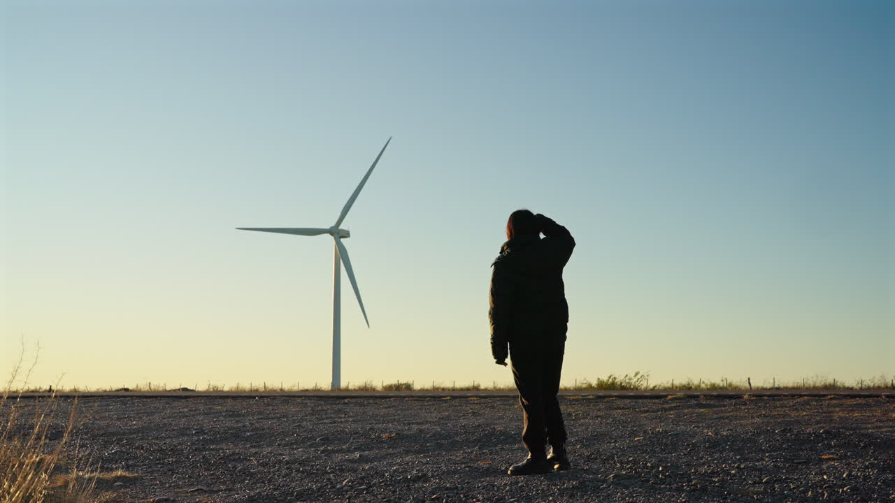 A lone person observes wind turbines spinning beneath a bright, clear blue sky