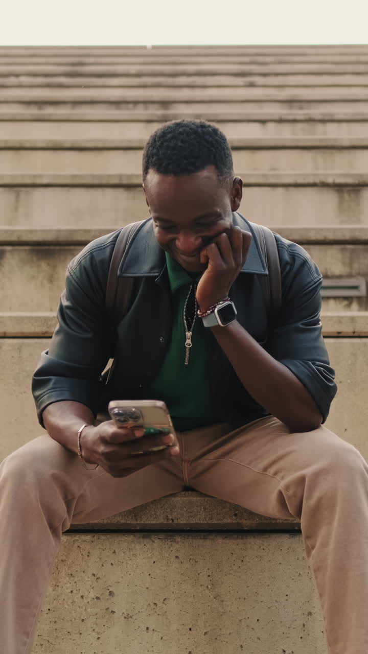 Young Black Man Sitting on Steps and Laughing While Using Smartphone